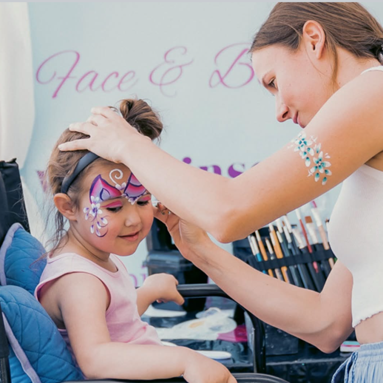 Kinderschminkteam Pinserella Eine Frau malt einem kleinen Mädchen bunte Gesichtsmalerei auf.