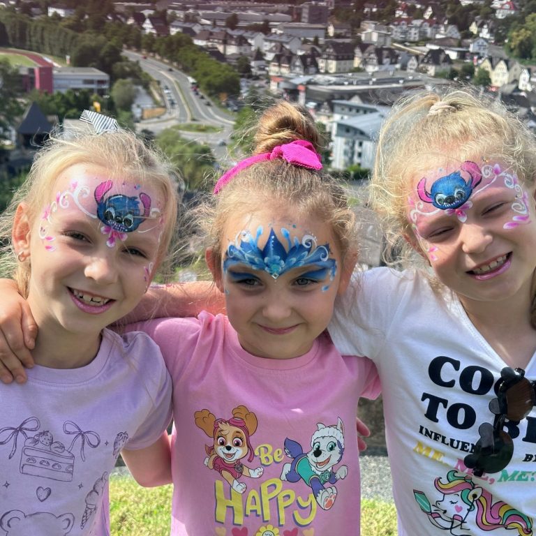 150 Jahre Wilhelmsturm Drei Mädchen mit bunten Gesichtsmalungen lächeln vor einer Stadtlandschaft.