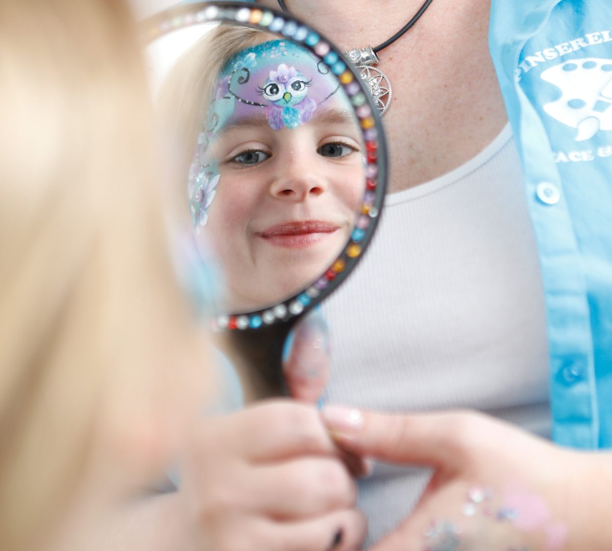Magische Momente * Kinderschminken mit Pinserella Mädchen lächelt in einen Spiegel, mit bunter Gesichtsbemalung.